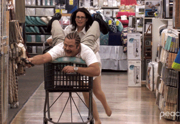 gif of Two characters from &quot;The Office&quot; are playfully engaging with one person pushing the other in a shopping cart