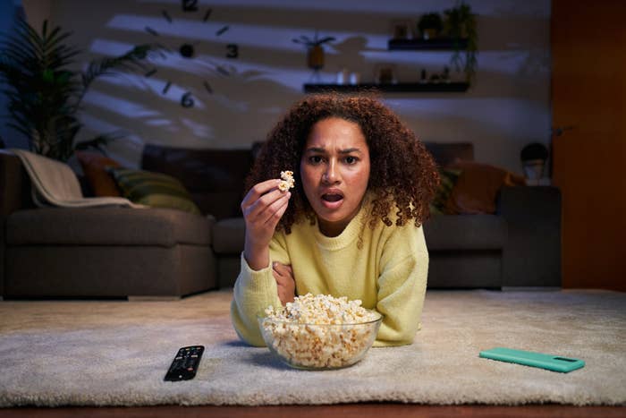 Woman watches TV with surprise, holding popcorn, remote and phone nearby