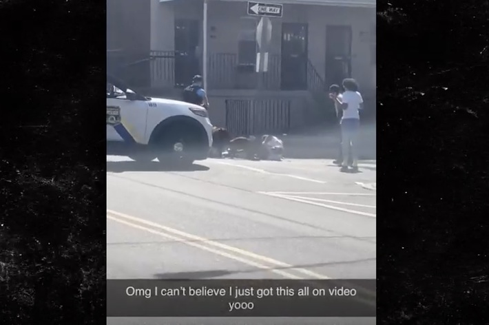 A person is filming a police incident involving an officer and a civilian on a street with bystanders watching