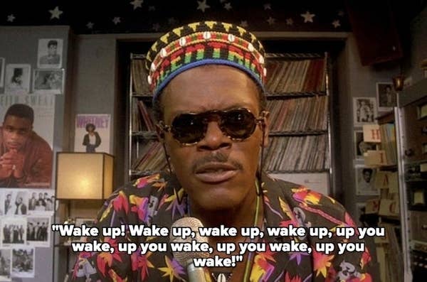 Samuel L. Jackson is wearing a colorful shirt and a patterned hat, speaking into a microphone in a radio studio. Text on image: "Wake up! Wake up, wake up, wake up, up you wake, up you wake, up you wake, up you wake!"