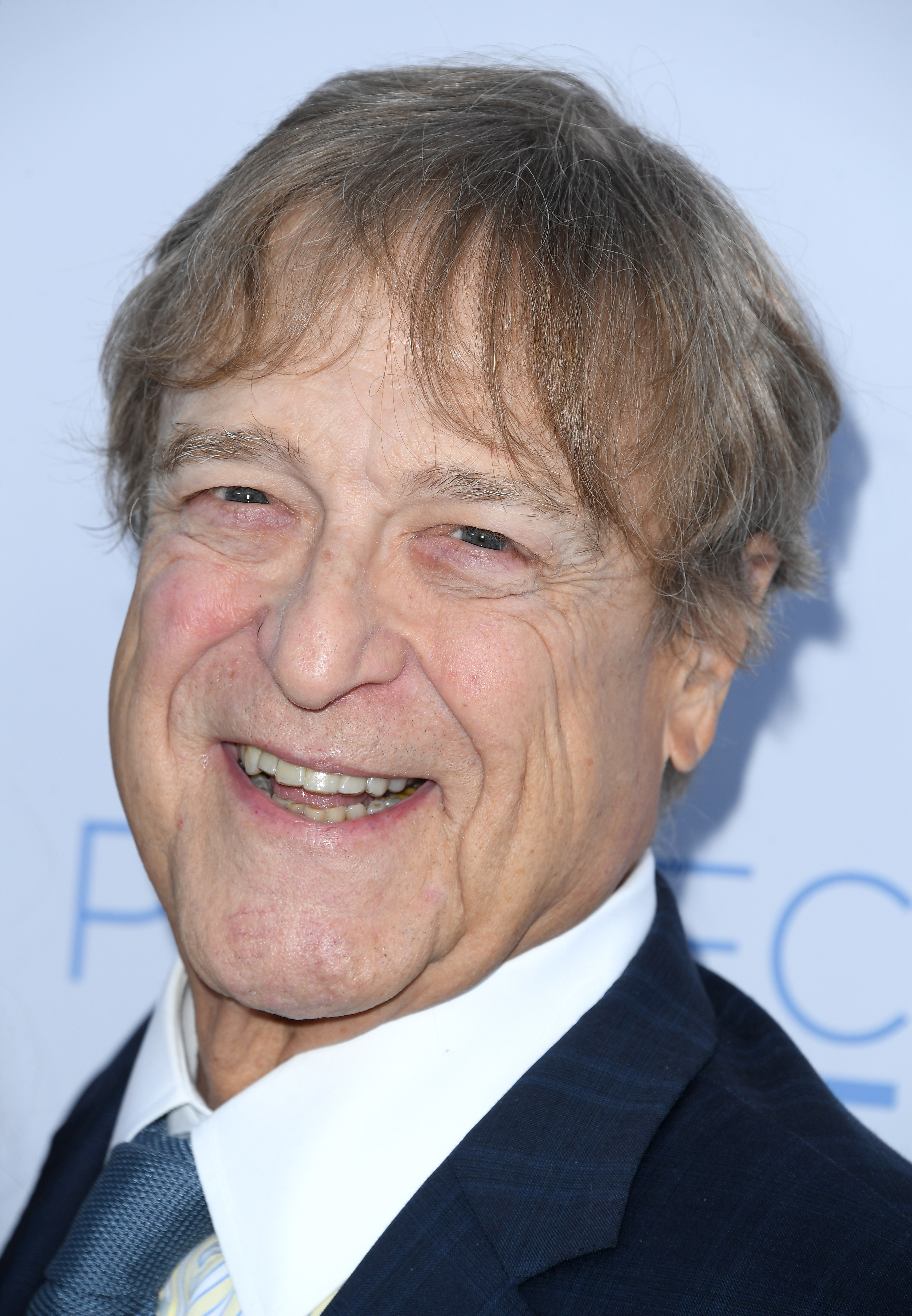 A man with light brown hair, wearing a dark suit and tie, smiles at the camera