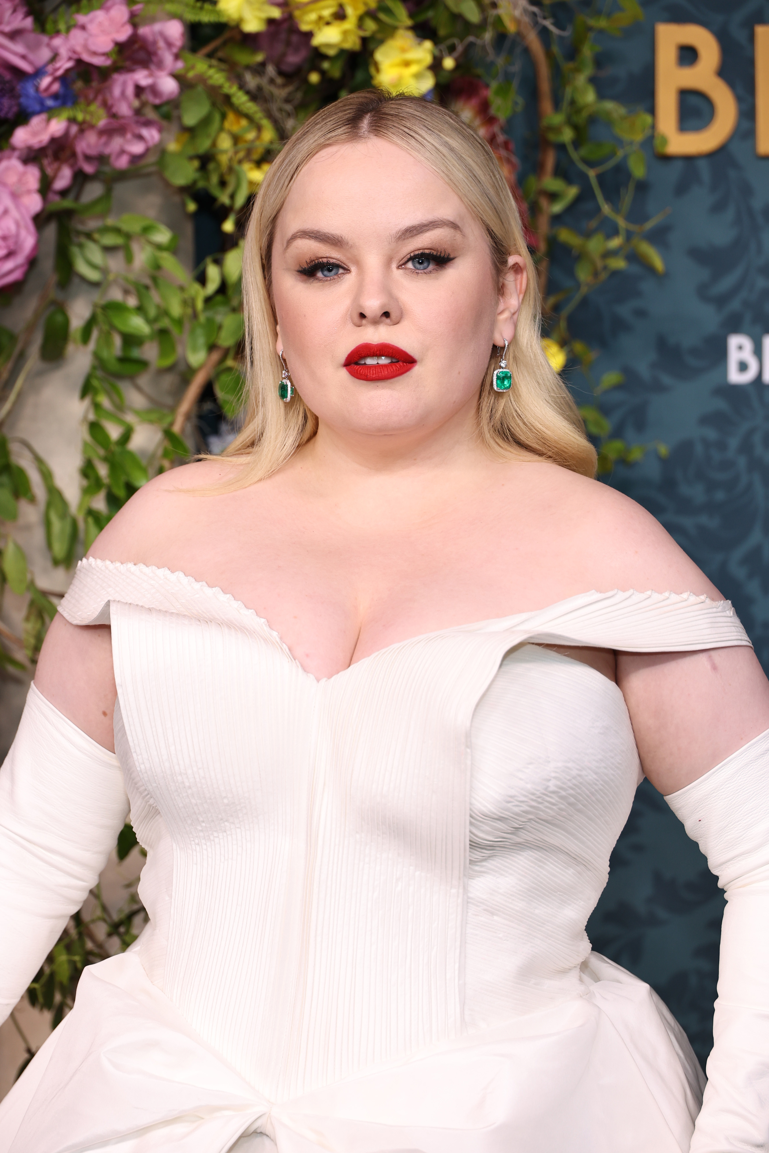 Nicola Coughlan in an off-the-shoulder white gown with statement earrings, posing in front of a floral backdrop at a Bridgerton event