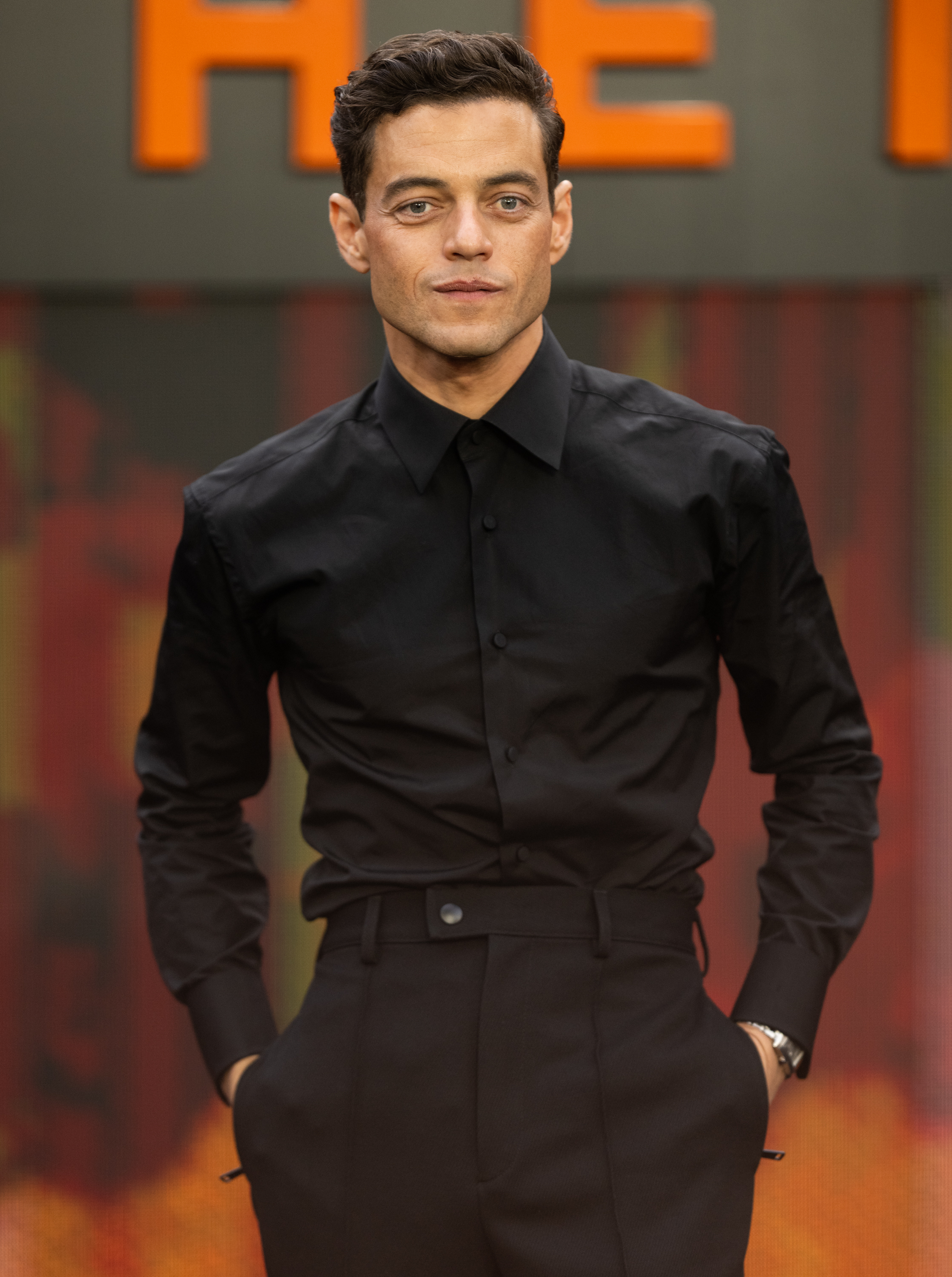 Rami Malek stands on a red carpet, wearing a sleek, black button-up shirt and high-waisted black trousers