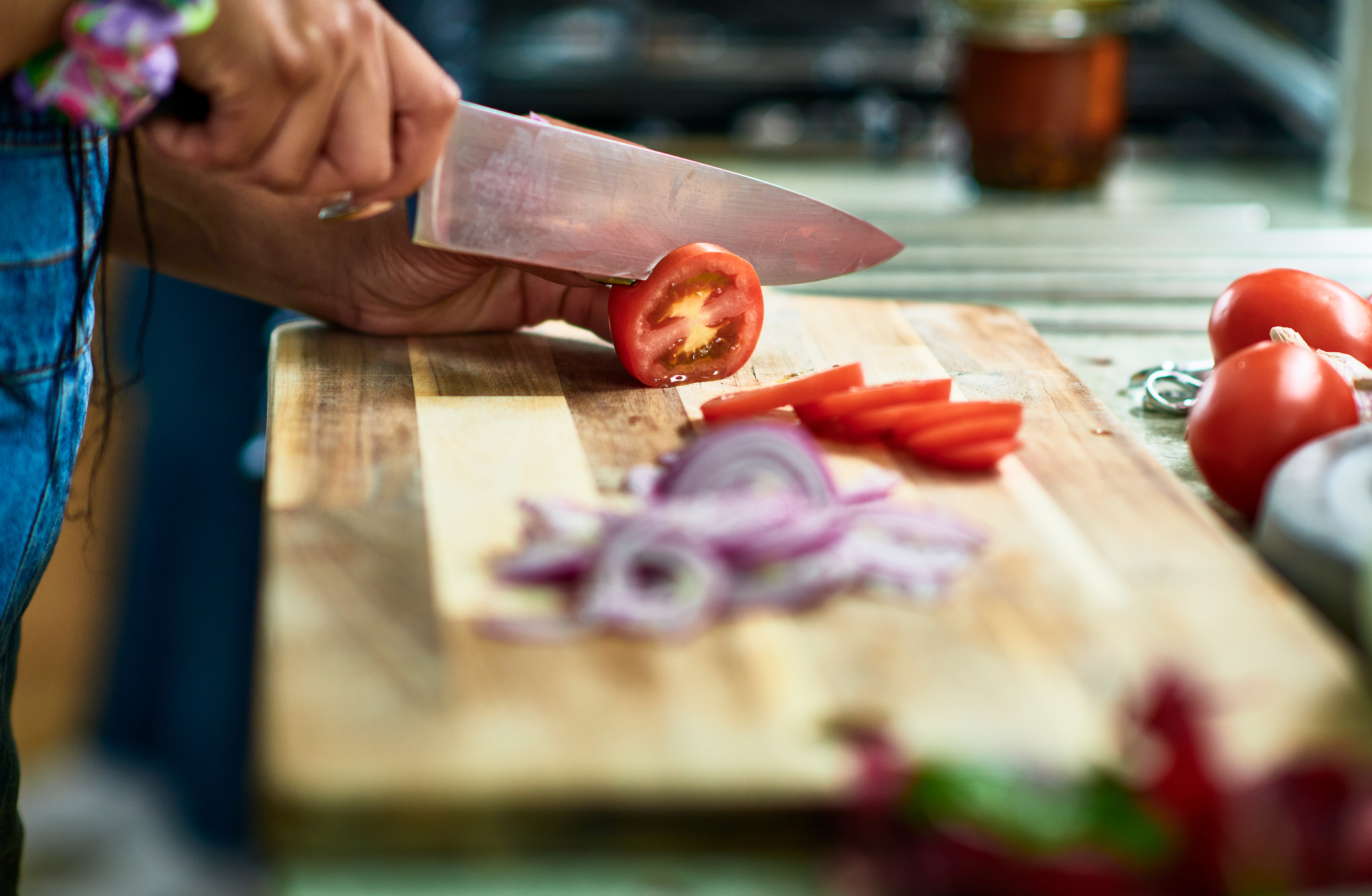 Close-up of a person slicing a tomato on a wooden cutting board, with sliced red onions and more tomatoes nearby in a kitchen setting