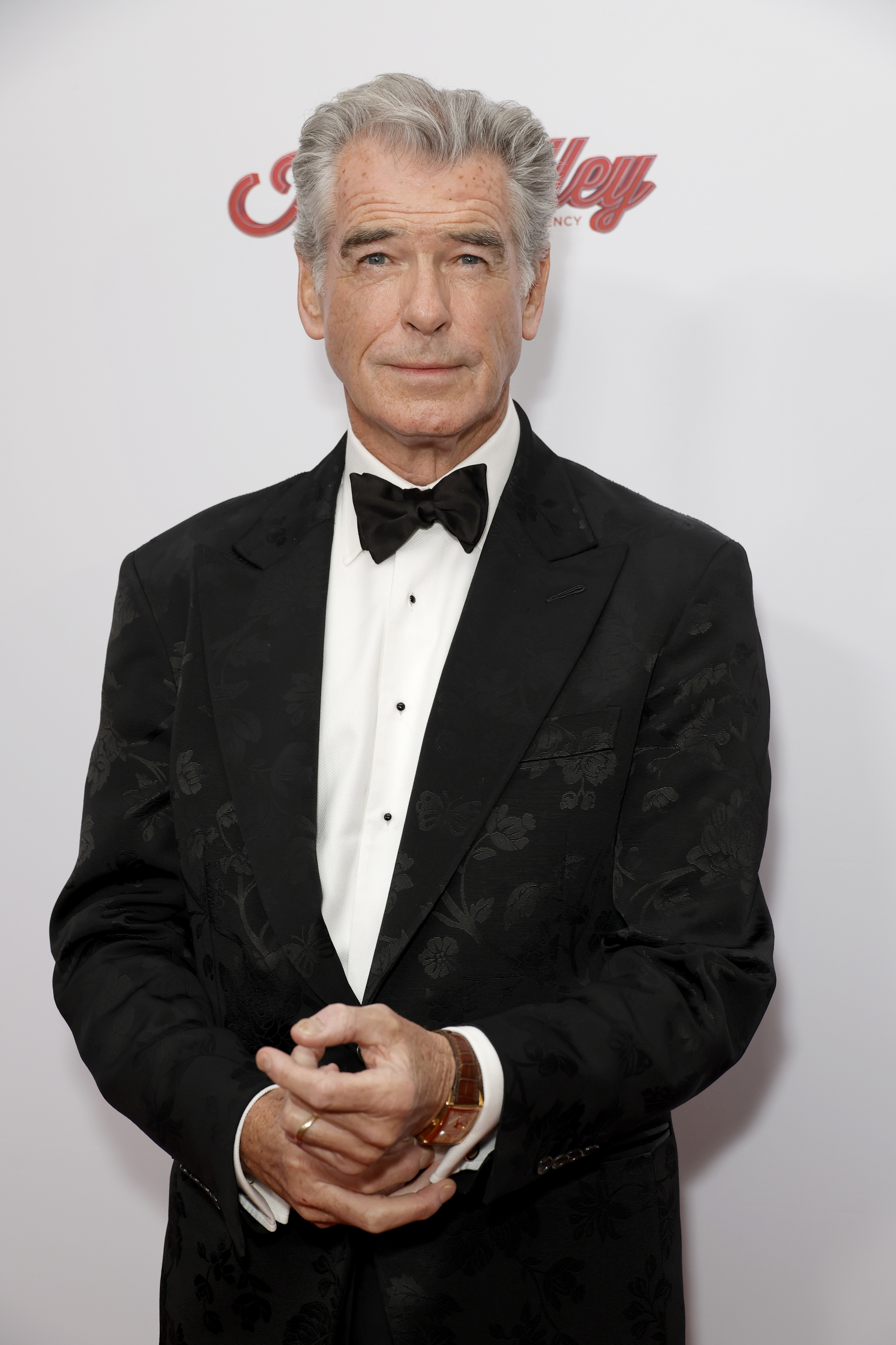 Pierce Brosnan wearing a black suit and bow tie, standing in front of a backdrop at a red carpet event