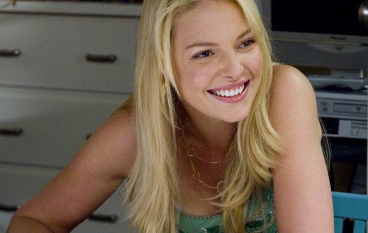 Katherine Heigl smiling while seated at a desk, wearing a casual sleeveless top