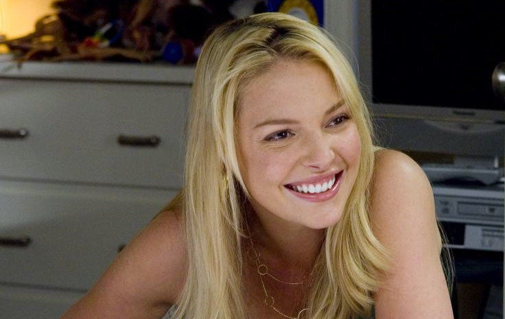 Katherine Heigl smiling while seated at a desk, wearing a casual sleeveless top