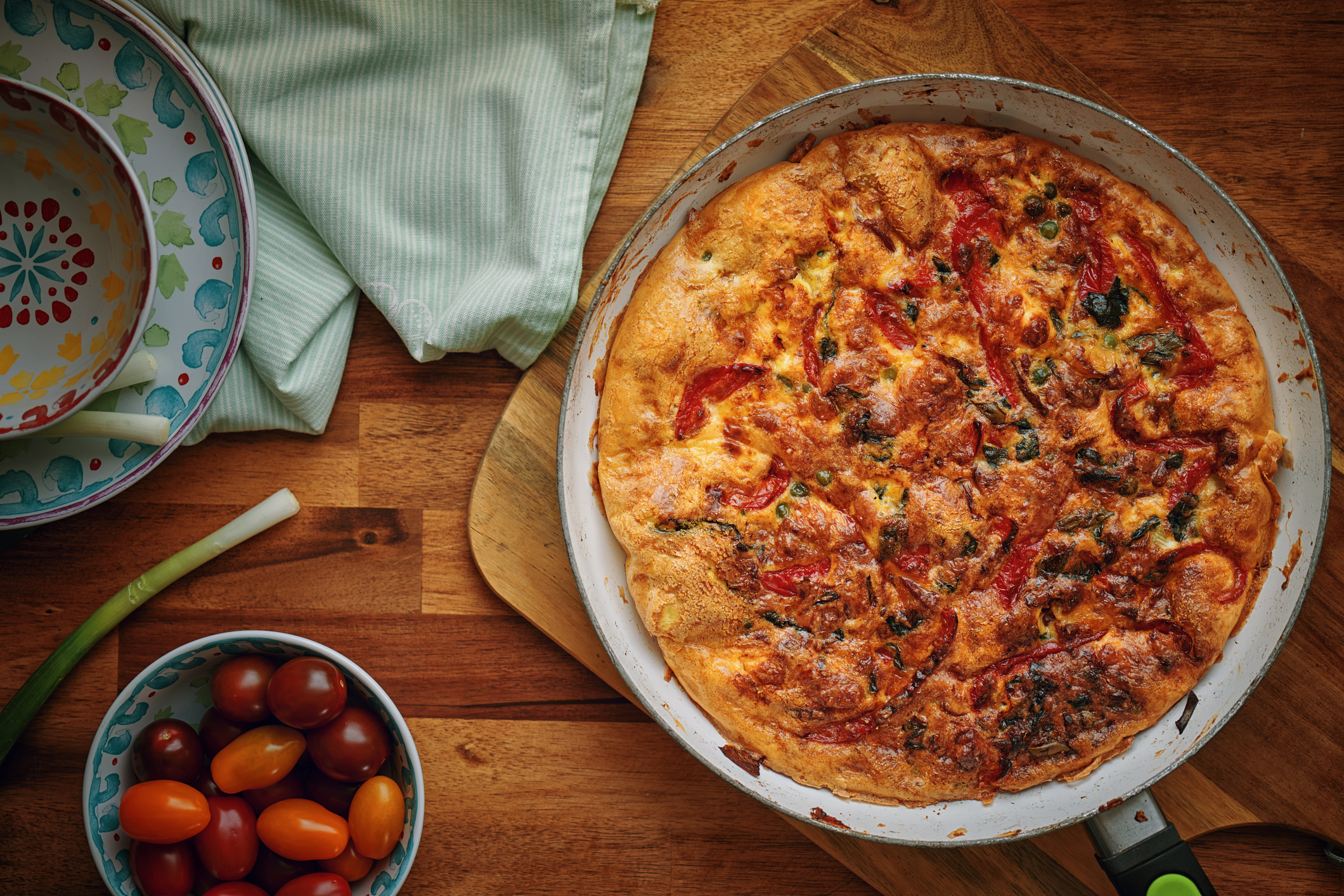 Top view of a freshly cooked frittata in a skillet on a wooden table with a bowl of cherry tomatoes, a spring onion, and a colorful ceramic plate