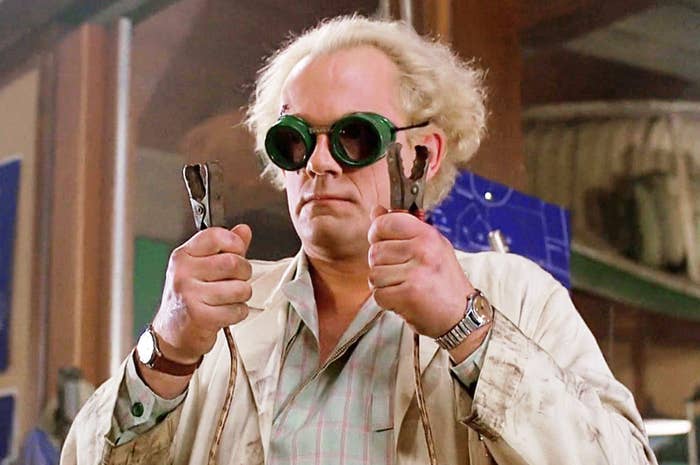 Christopher Lloyd as Doc Brown in eccentric scientist attire, holding two jumper cables with a focused expression, from the movie &quot;Back to the Future.&quot;