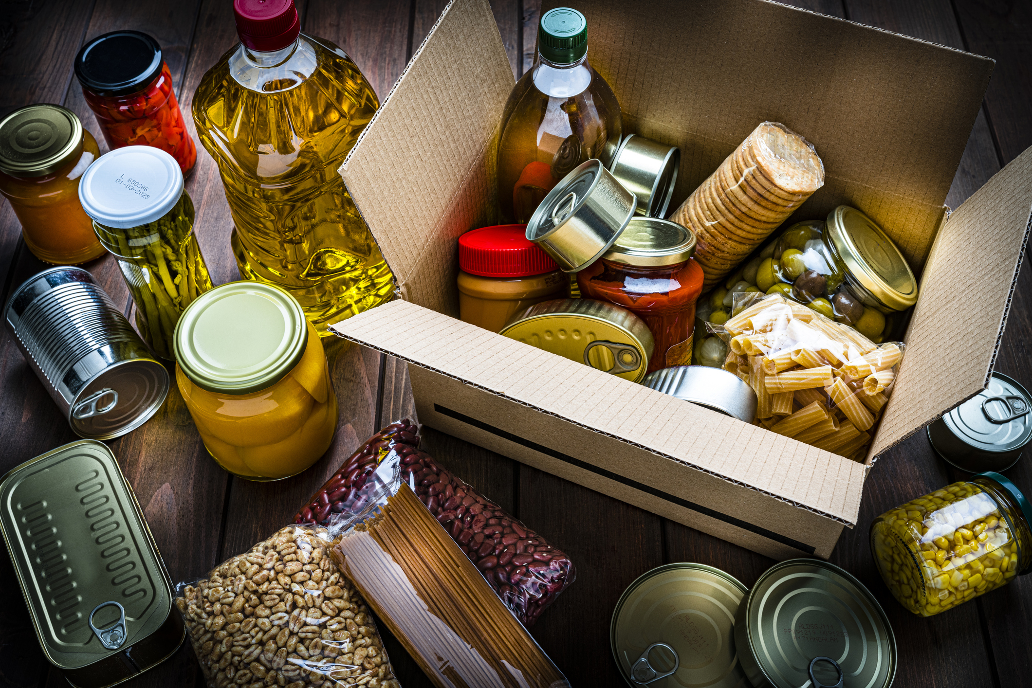 Various food items, including jars, cans, pasta, and oil, are placed around and inside an open cardboard box on a wooden surface