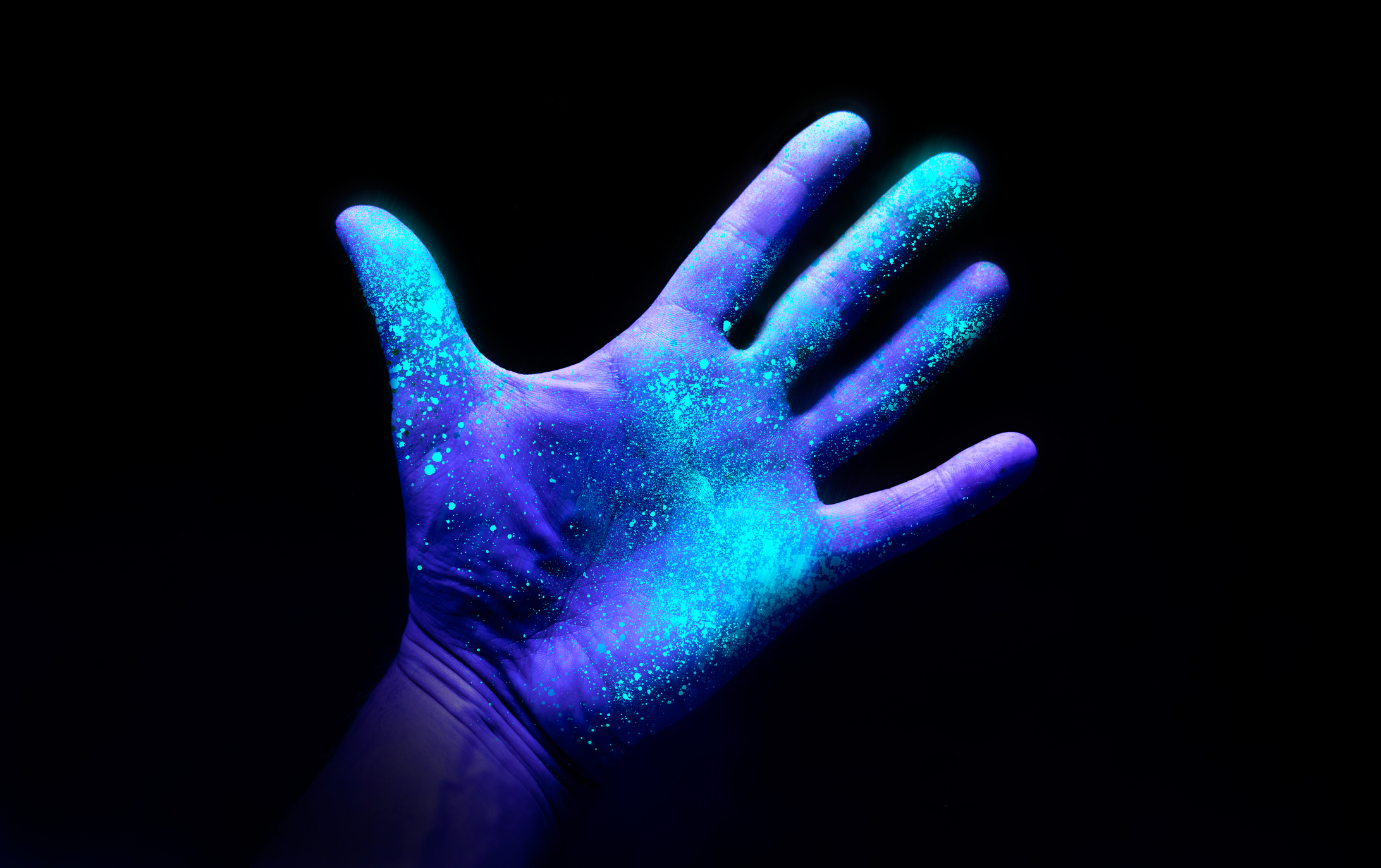 A close-up of a hand covered in neon, glowing specks under UV light against a dark background