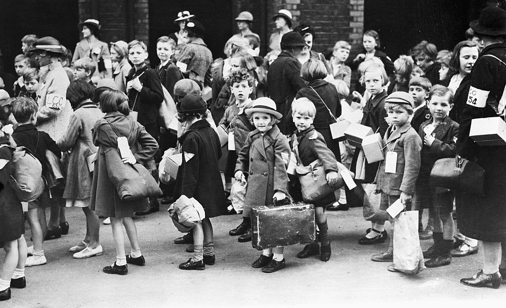 A crowd of children with luggage and name tags, accompanied by adults, are gathered outside a brick building during World War II evacuation