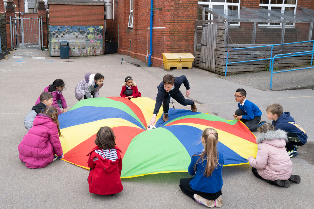 Children play a parachute game in a schoolyard, each holding an edge while one child in the center touches the fabric with a ball on the top