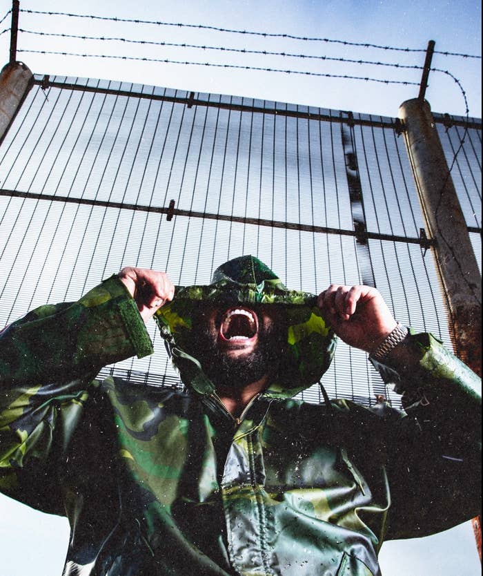 Person wearing a camo raincoat and hat, holding the hat&#x27;s brim while screaming or shouting, posed in front of a tall fence with barbed wire on top
