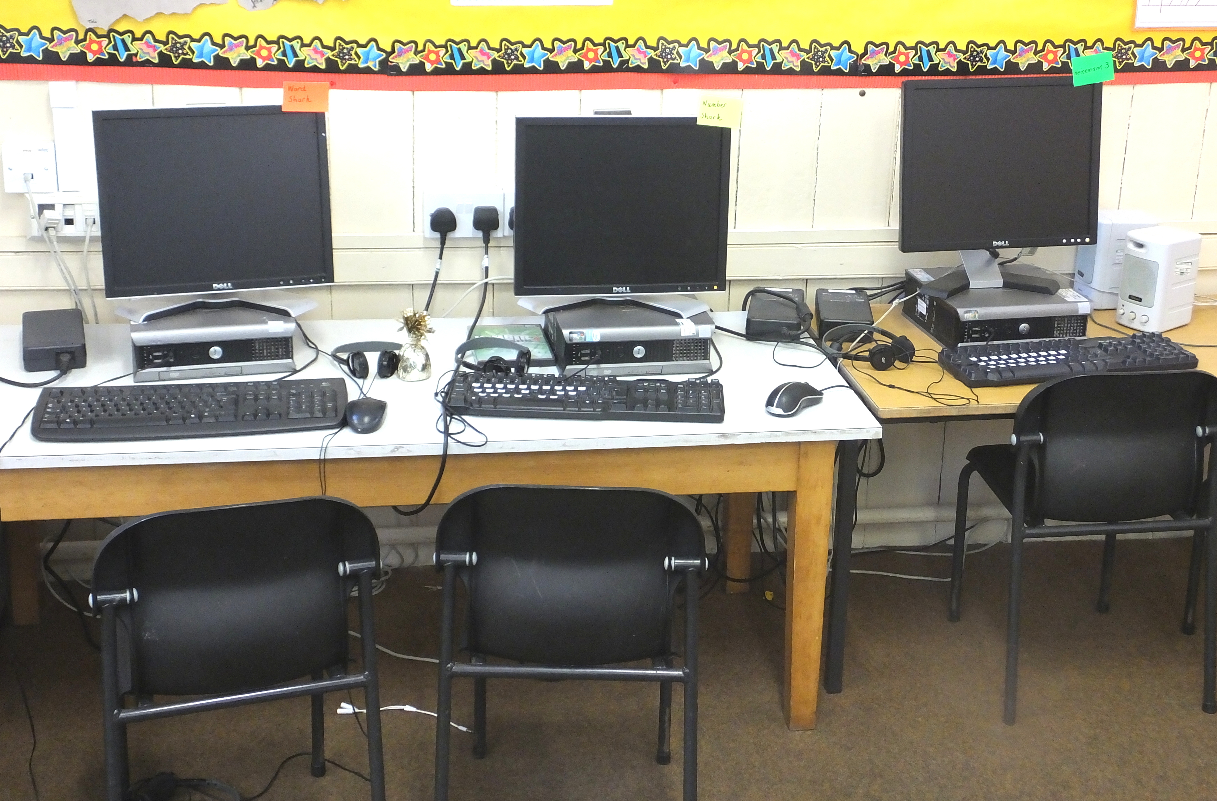 Three computer workstations with monitors and keyboards are set up on a desk, accompanied by three black chairs. A decorative border with stars is above the desk