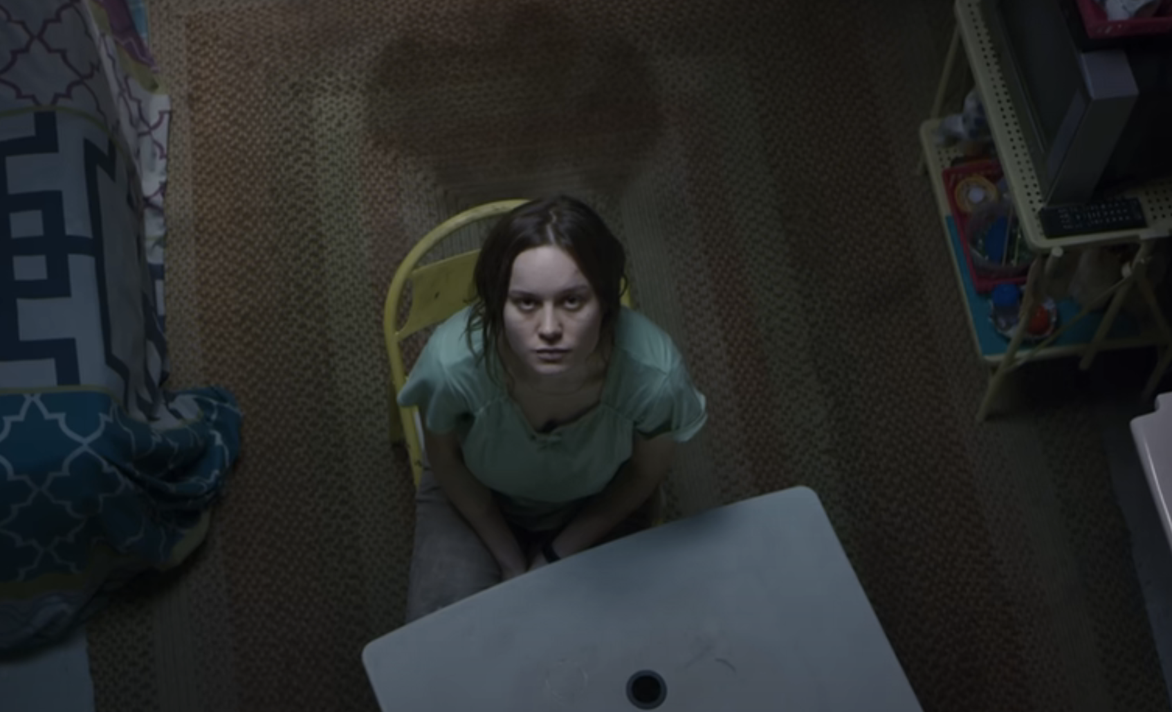 Brie Larson sitting on a chair in a sparse room, looking up towards the camera with a serious expression in a scene from Room