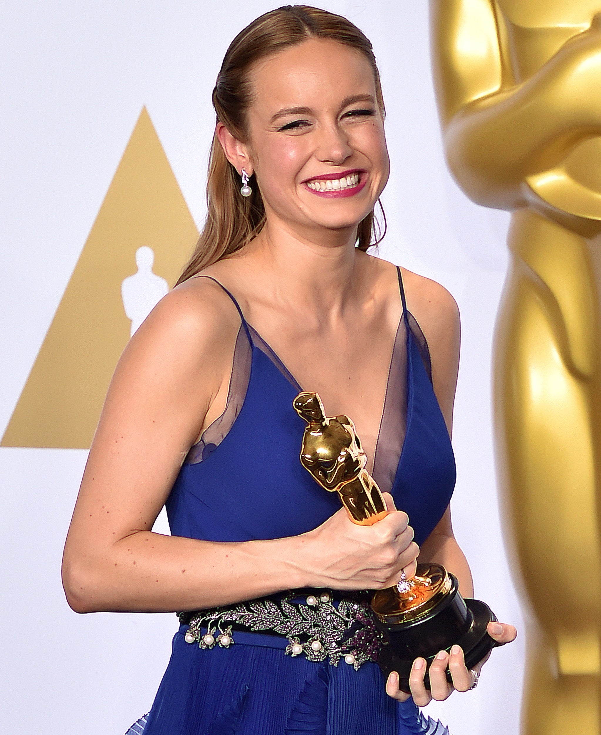 Brie Larson, smiling and holding her Oscar trophy, in a stylish sleeveless gown with floral details