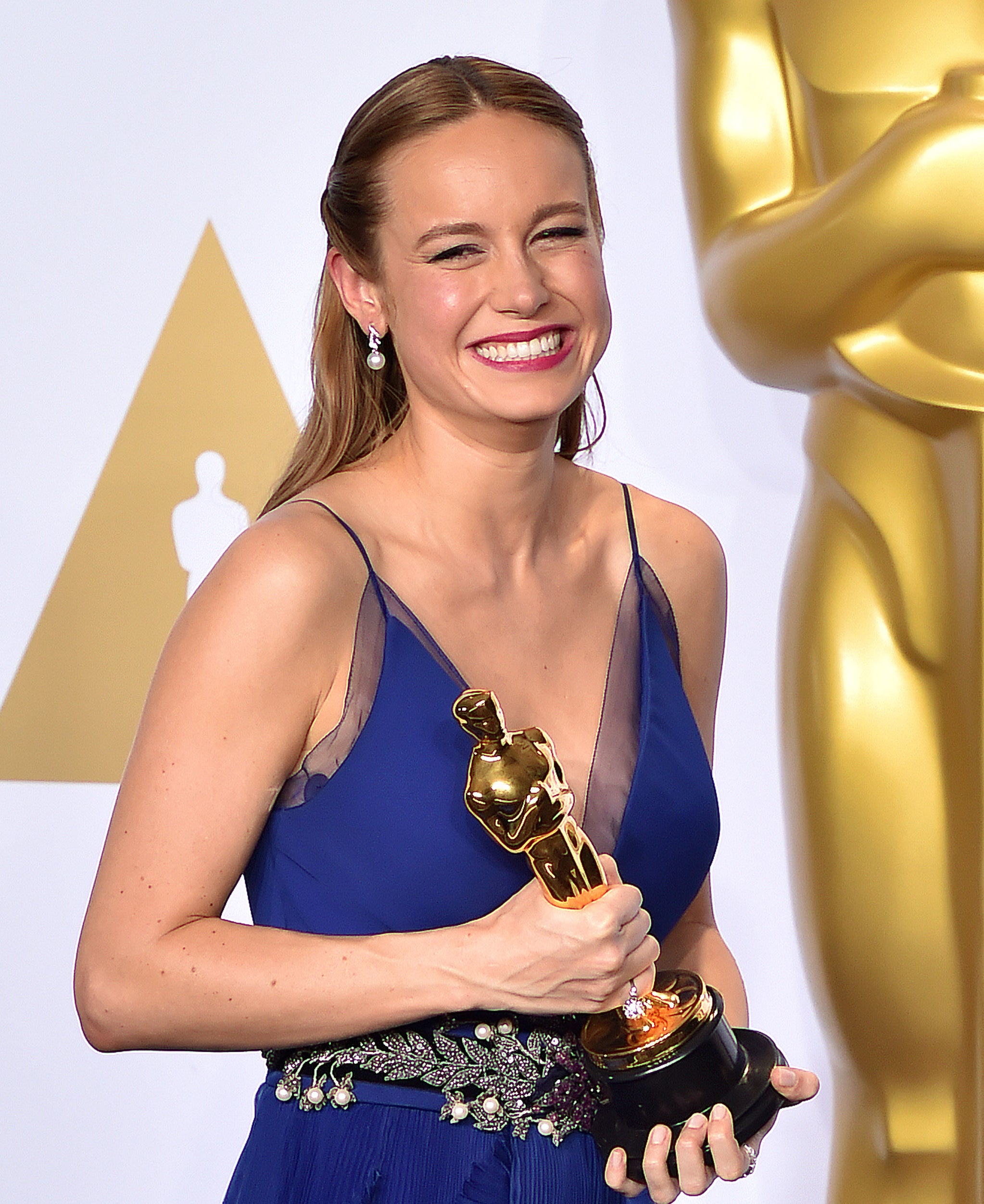 Brie Larson, smiling, holding an Oscar trophy, in a stylish sleeveless gown with floral details, standing in front of an Oscar statue