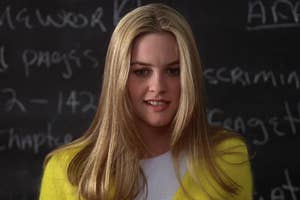 Alicia Silverstone in a classroom scene from the movie "Clueless," wearing a white top and a yellow cardigan. The blackboard behind her has partially visible chalk writing