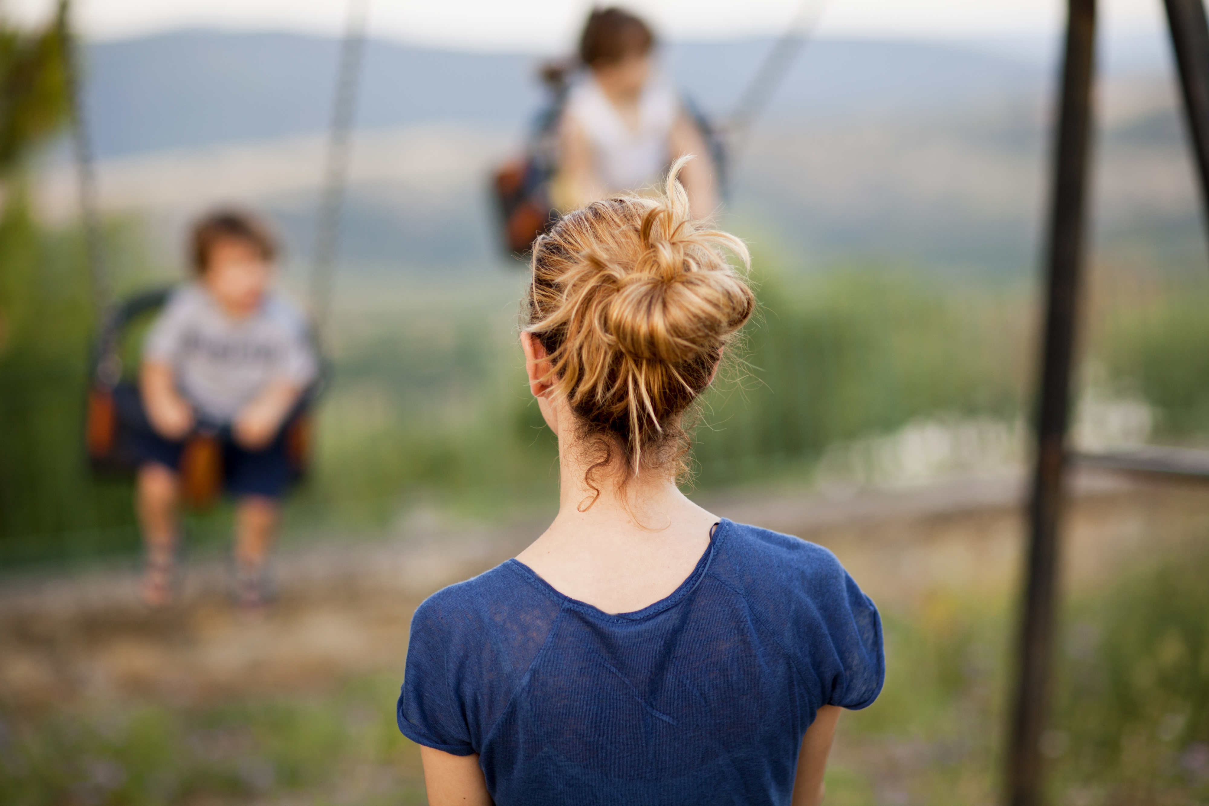 A woman with a bun hairstyle watches two children on swings at a playground