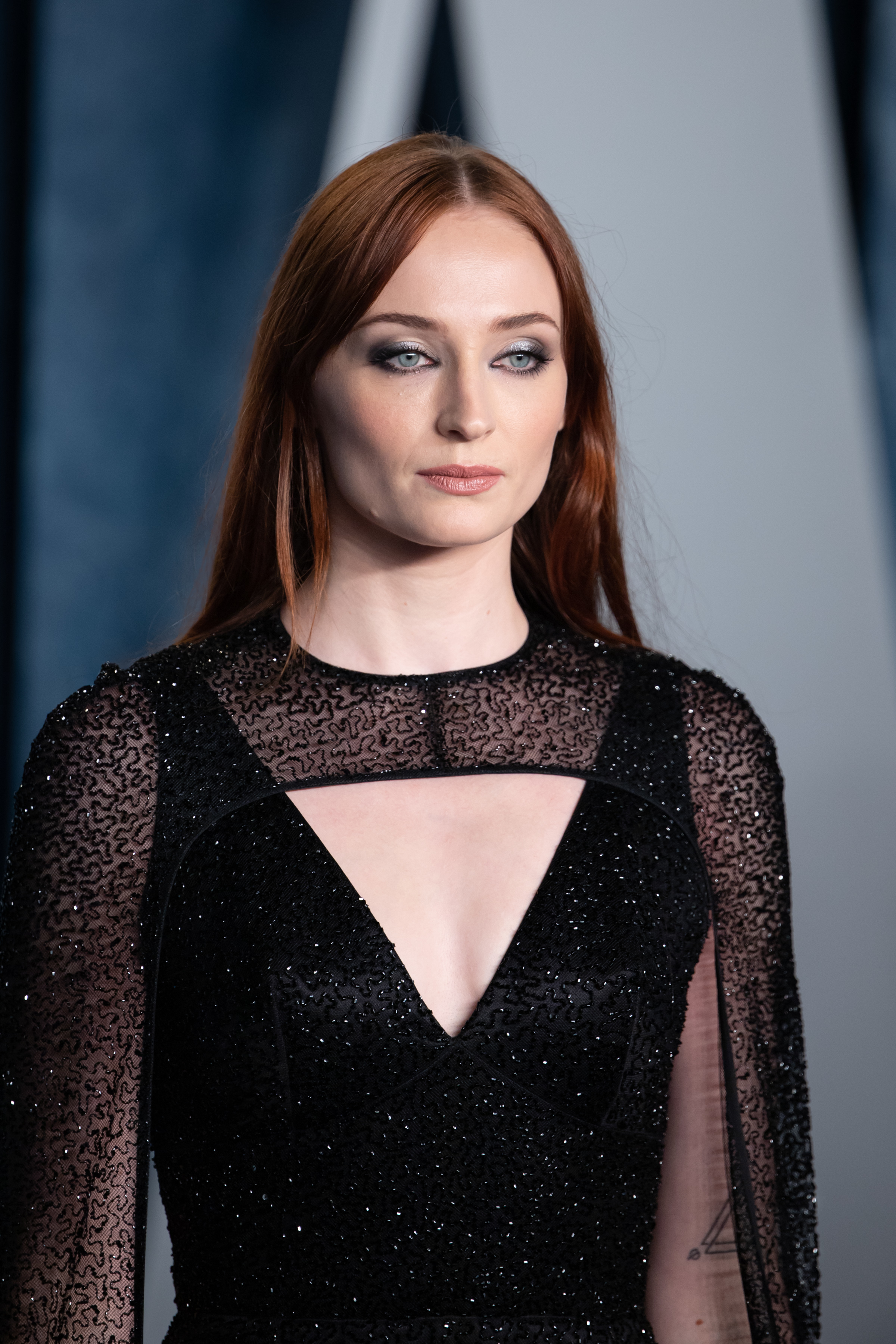 Sophie Turner at a formal event, wearing a black sequined gown with sheer sleeves and geometric cutout neckline