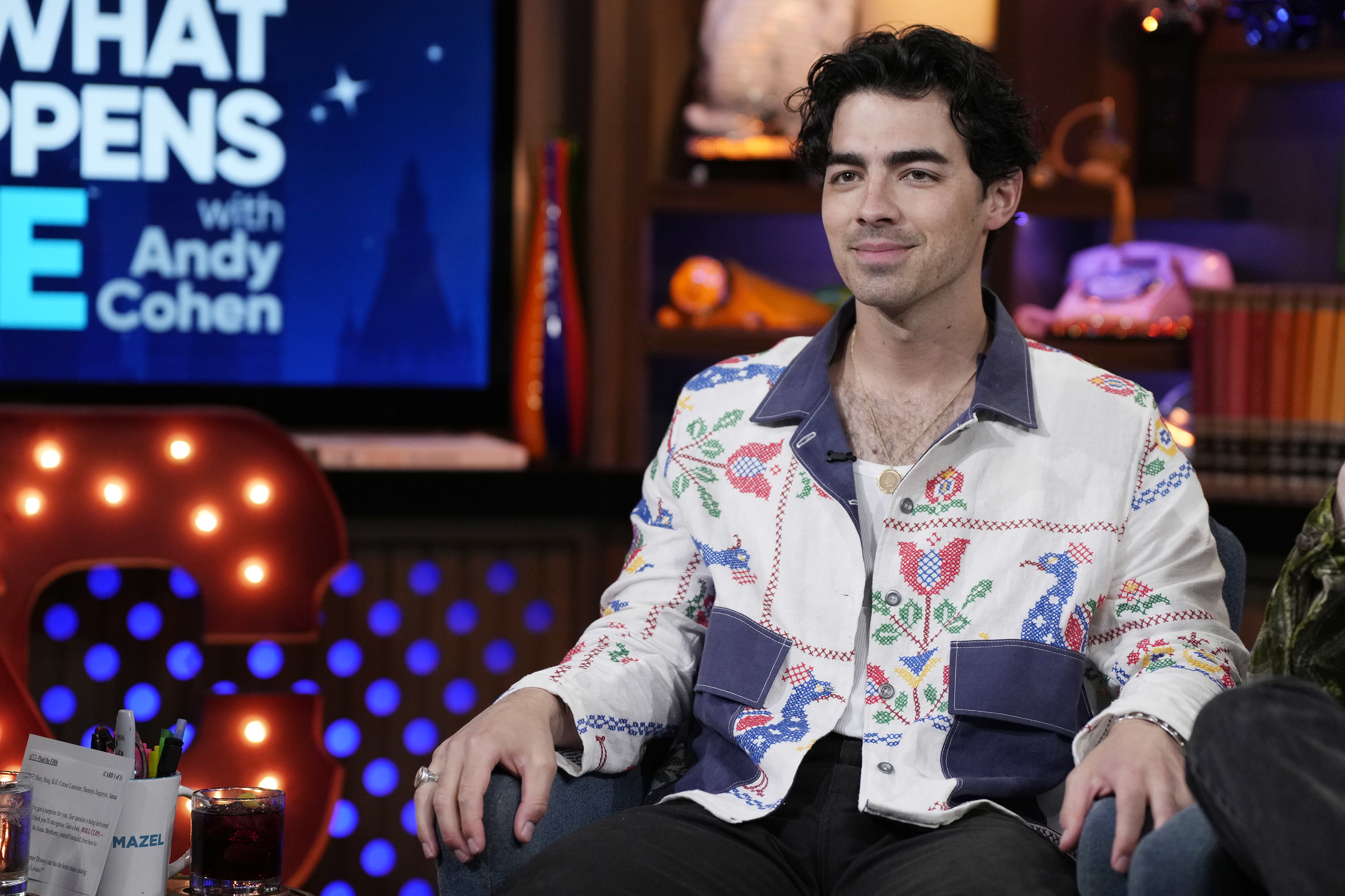 Joe Jonas, seated, wearing a white jacket with colorful floral embroidery, on the set of &quot;Watch What Happens Live with Andy Cohen.&quot;