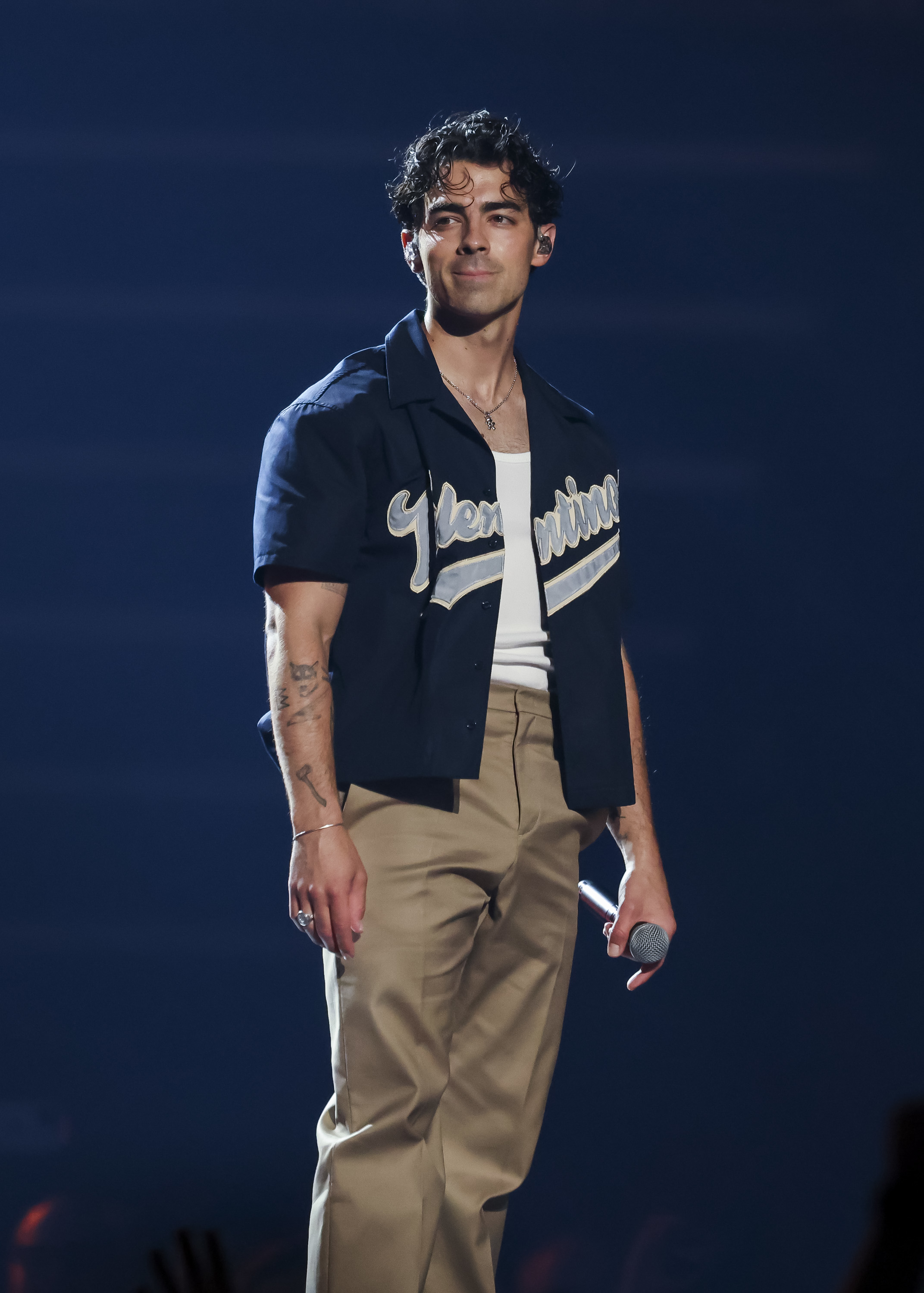 Joe Jonas stands on a stage, wearing a &quot;Youngstown&quot; shirt, tan pants, and holding a microphone