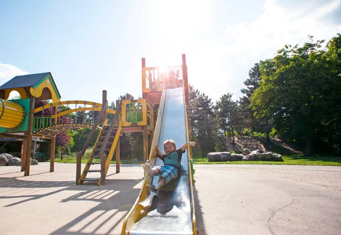 A child is joyfully sliding down a playground slide on a sunny day, with trees and playground structures in the background