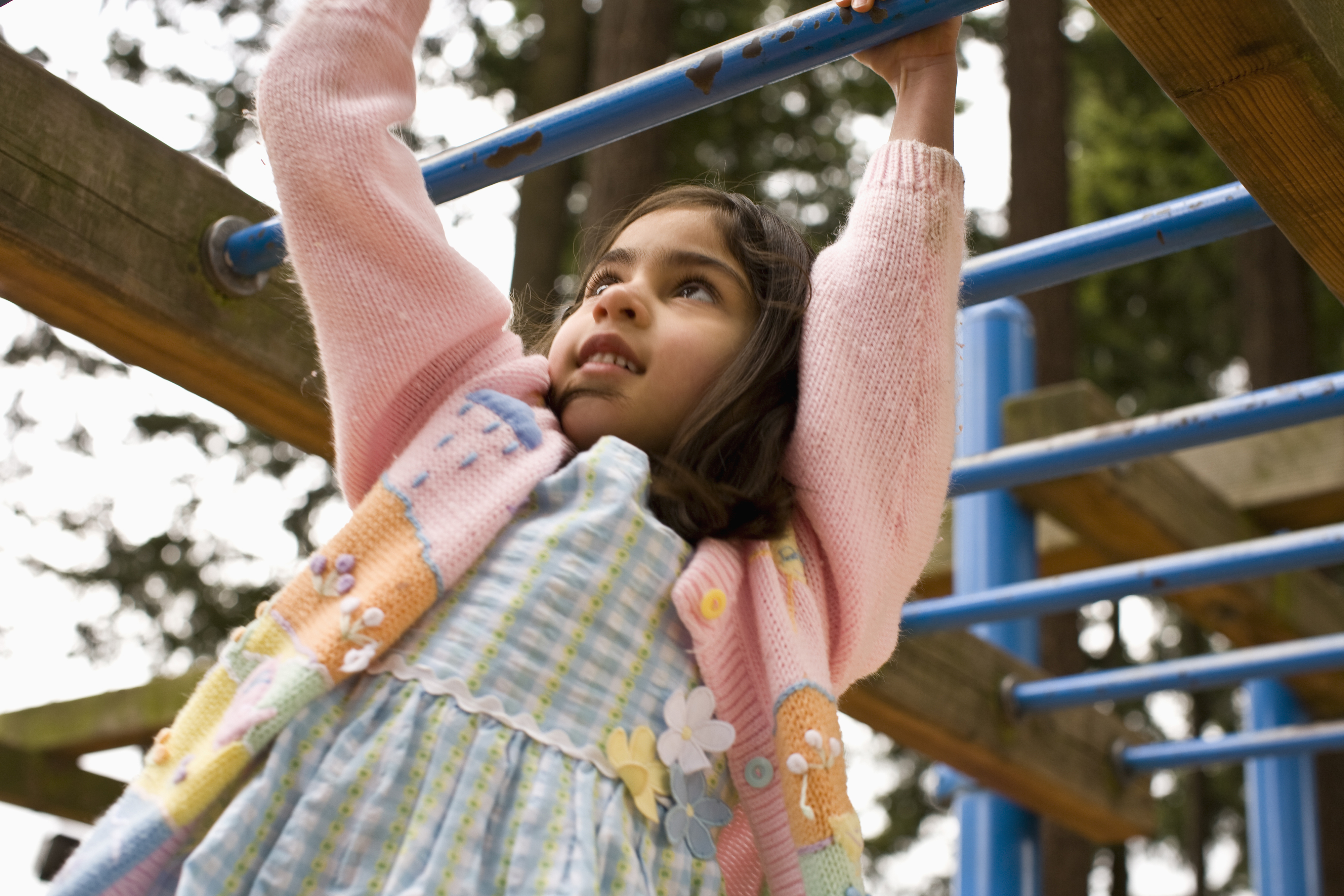 A young girl wearing a dress and cardigan hangs from a monkey bar in a playground, looking determined