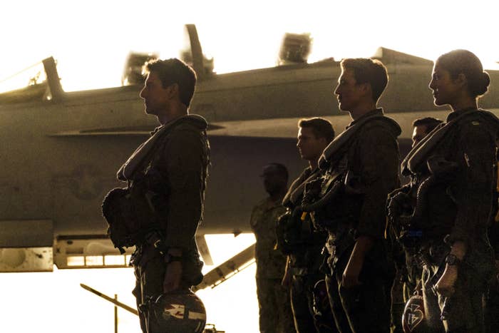 Tom Cruise standing in front of a fighter jet with Miles Teller, Glen Powell, and Monica Barbaro, all dressed in flight suits