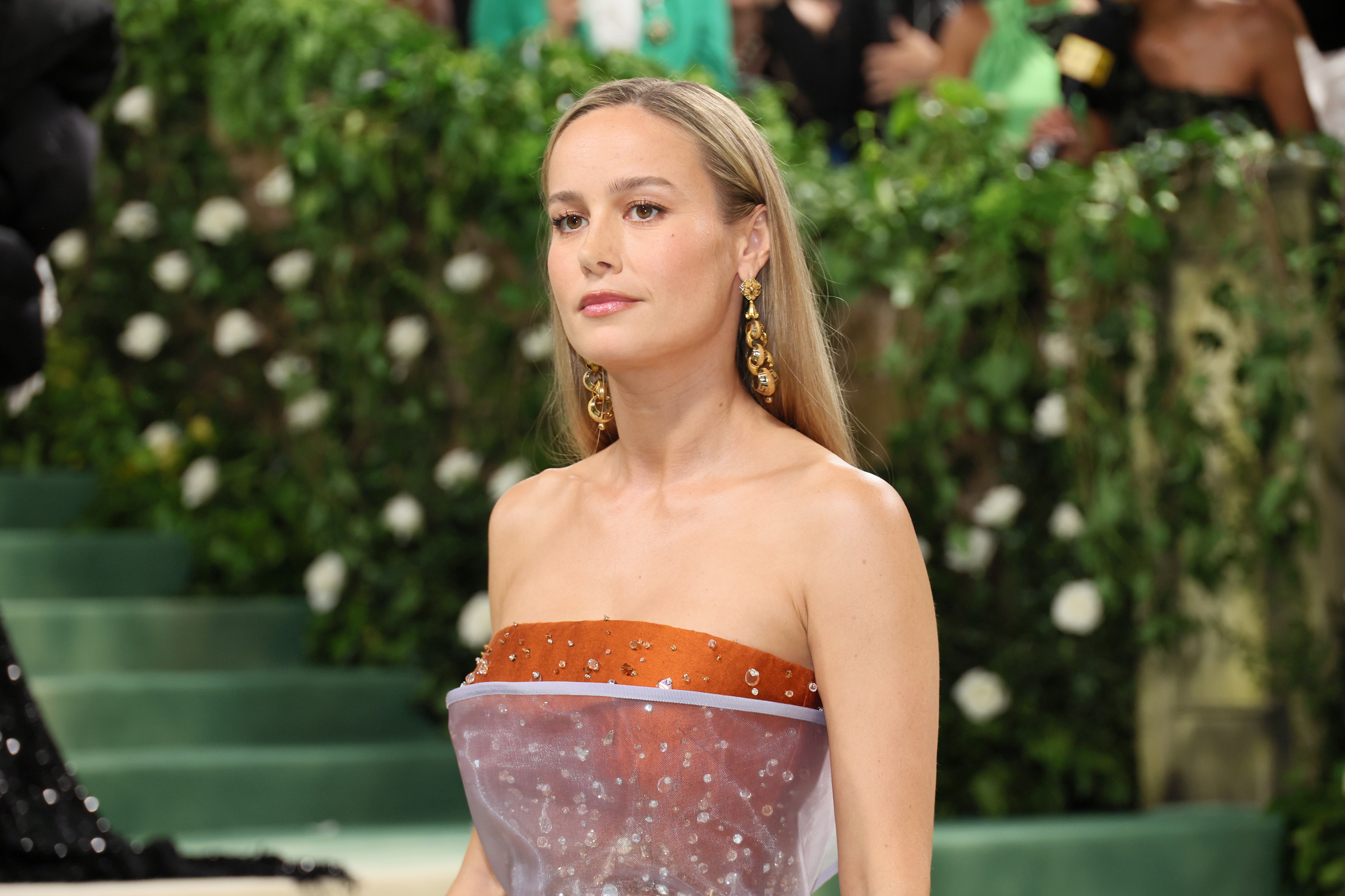 Brie Larson at an event in a strapless gown with gold earrings, standing in front of a greenery-decorated background