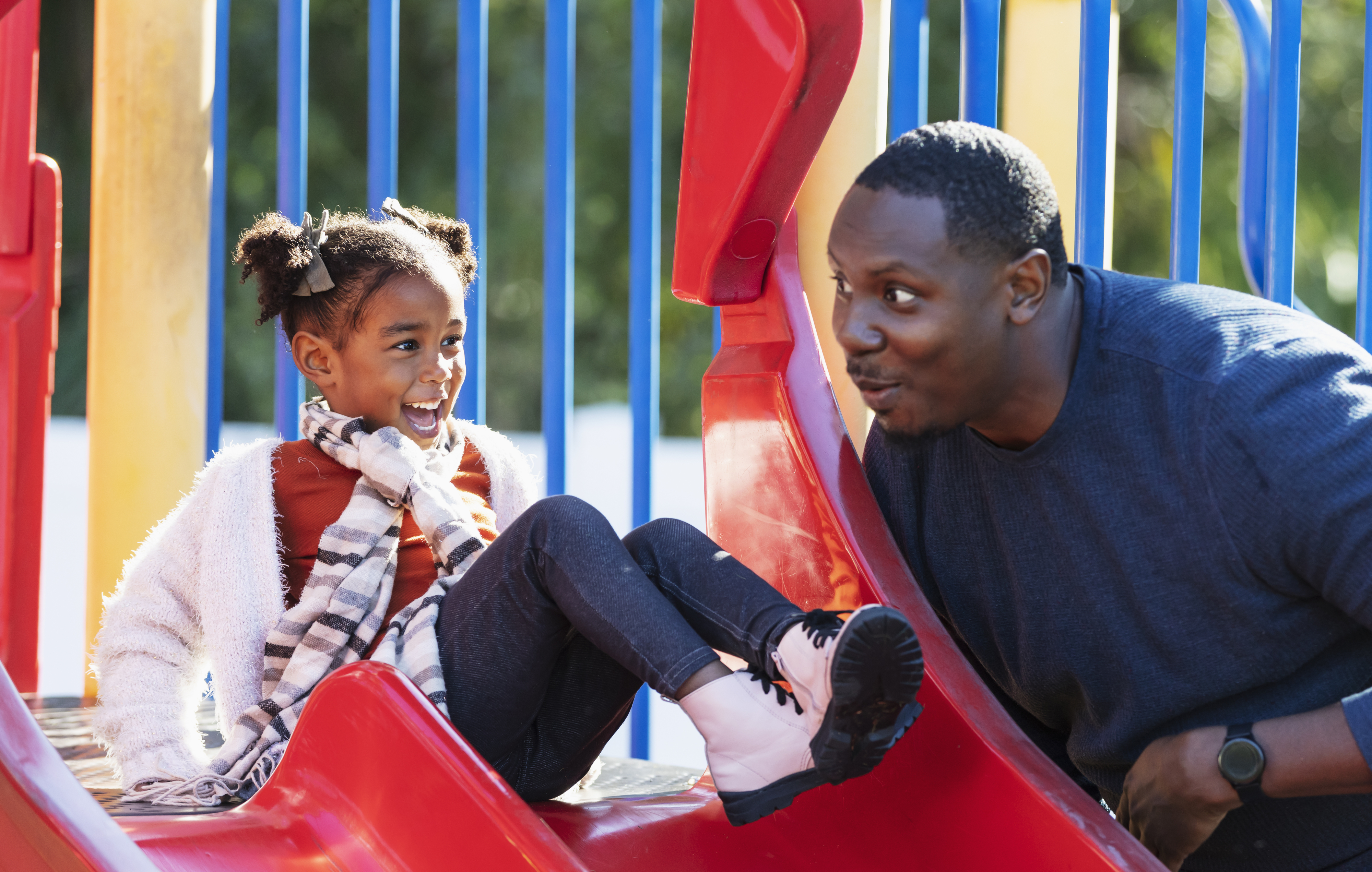A man in a blue sweater interacts playfully with a young girl sitting on a slide. Both are smiling and appear to be enjoying the moment