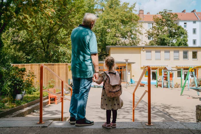 An older man and a young girl holding hands stand at the entrance of a playground, facing away from the camera. The girl is wearing a backpack