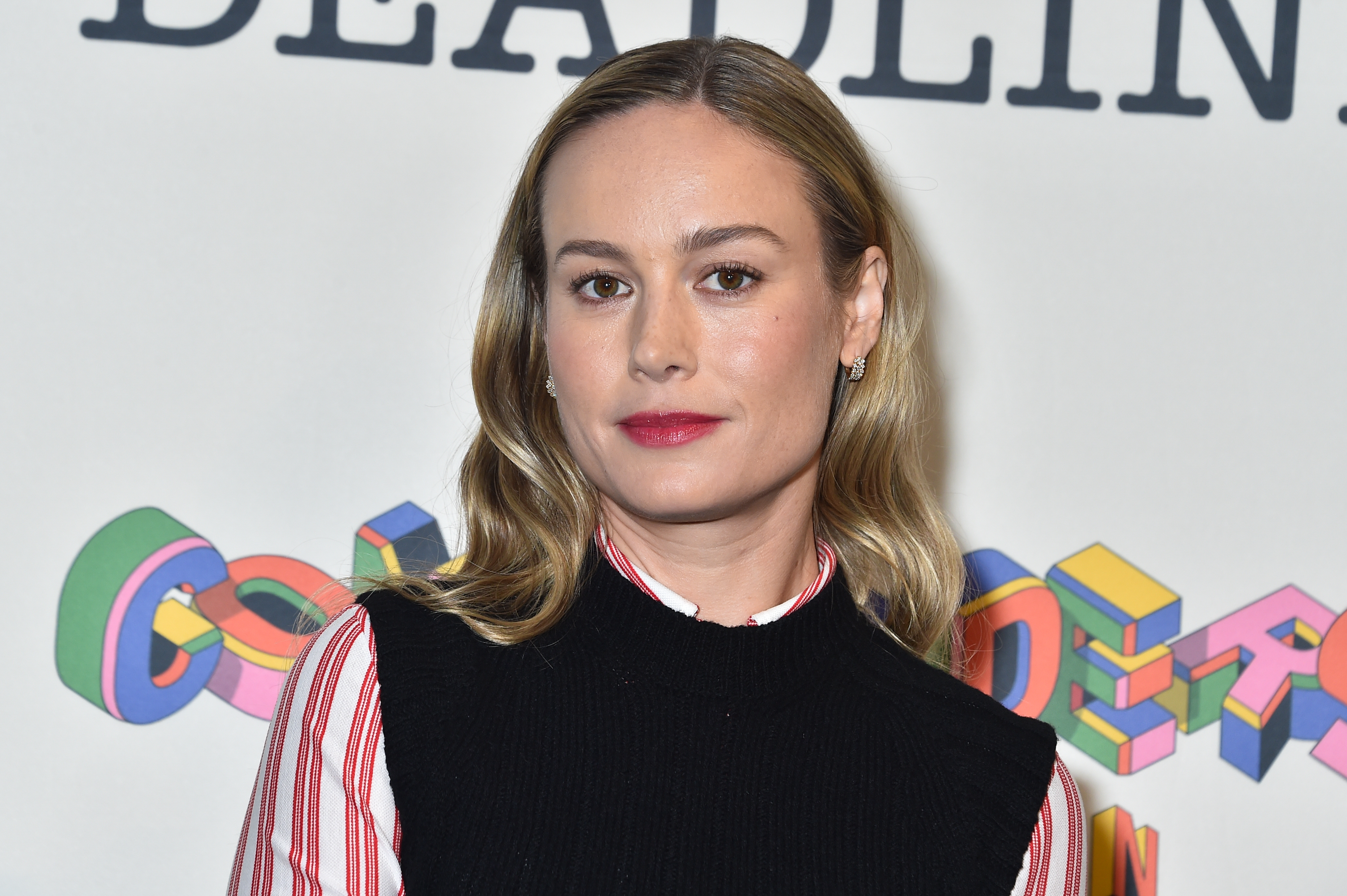 Brie Larson standing against a backdrop with text. She is wearing a black sleeveless dress over a red and white striped shirt