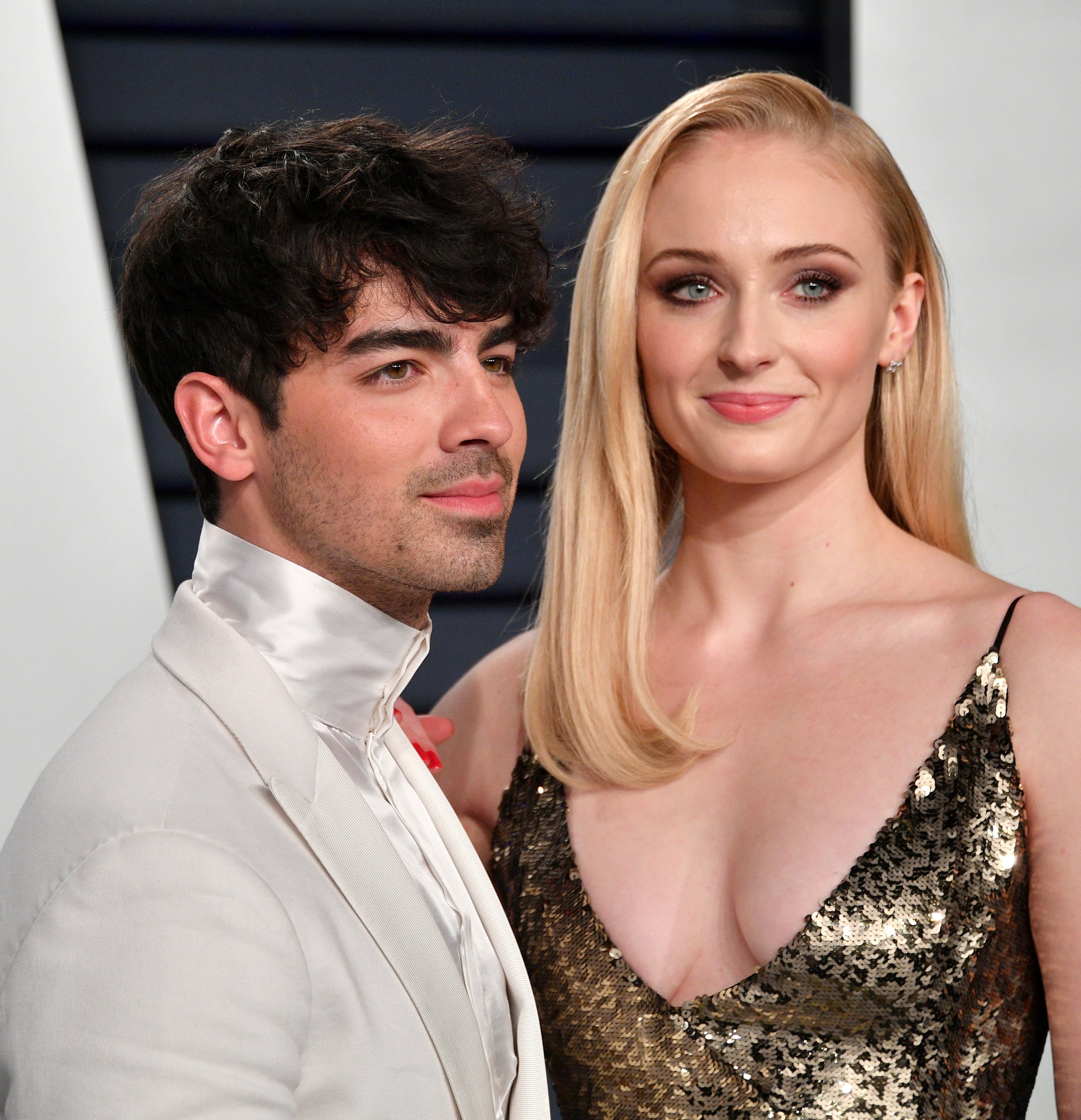 Joe Jonas in a white suit stands beside Sophie Turner in a sleeveless, sequined gown, posing for a photo