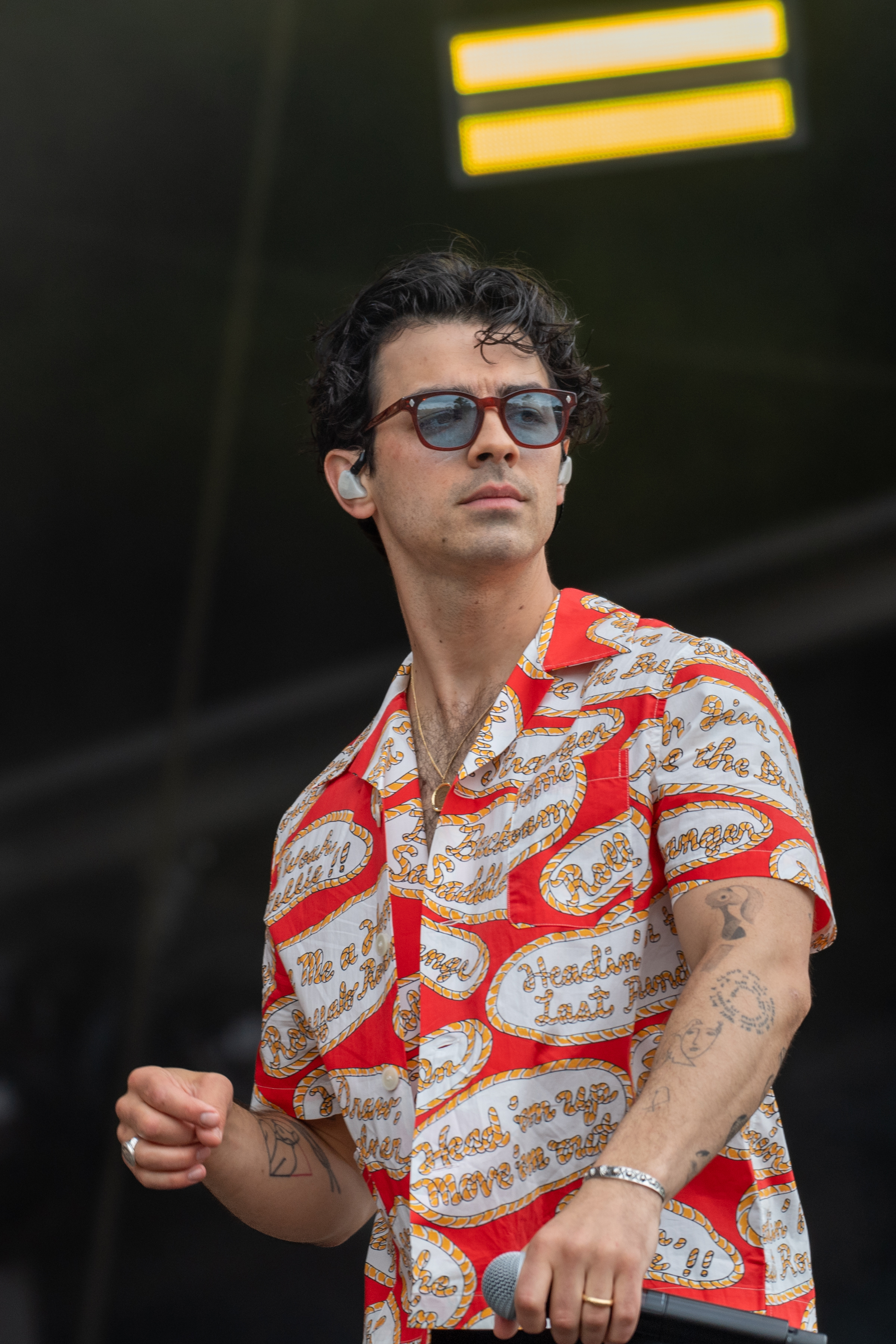 Joe Jonas wearing a patterned short-sleeve shirt and sunglasses performs on stage, holding a microphone