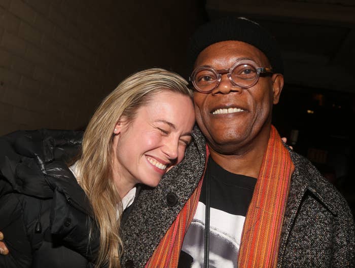 Brie Larson smiles with eyes closed while hugging Samuel L. Jackson, who is wearing a knit hat, glasses, and a patterned scarf indoors