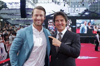 Glen Powell in a blue suit and Tom Cruise in a black suit pose and smile for photos on a red carpet at an event