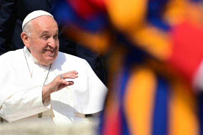 Pope Francis smiles and gestures while seated, with the blurred figure of a Swiss Guard in vibrant attire nearby