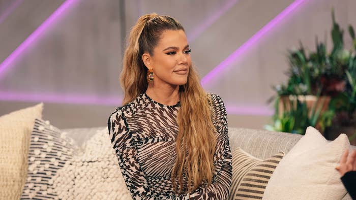 Khloé Kardashian in a patterned dress, sits on a sofa during an interview, smiling and looking to the side