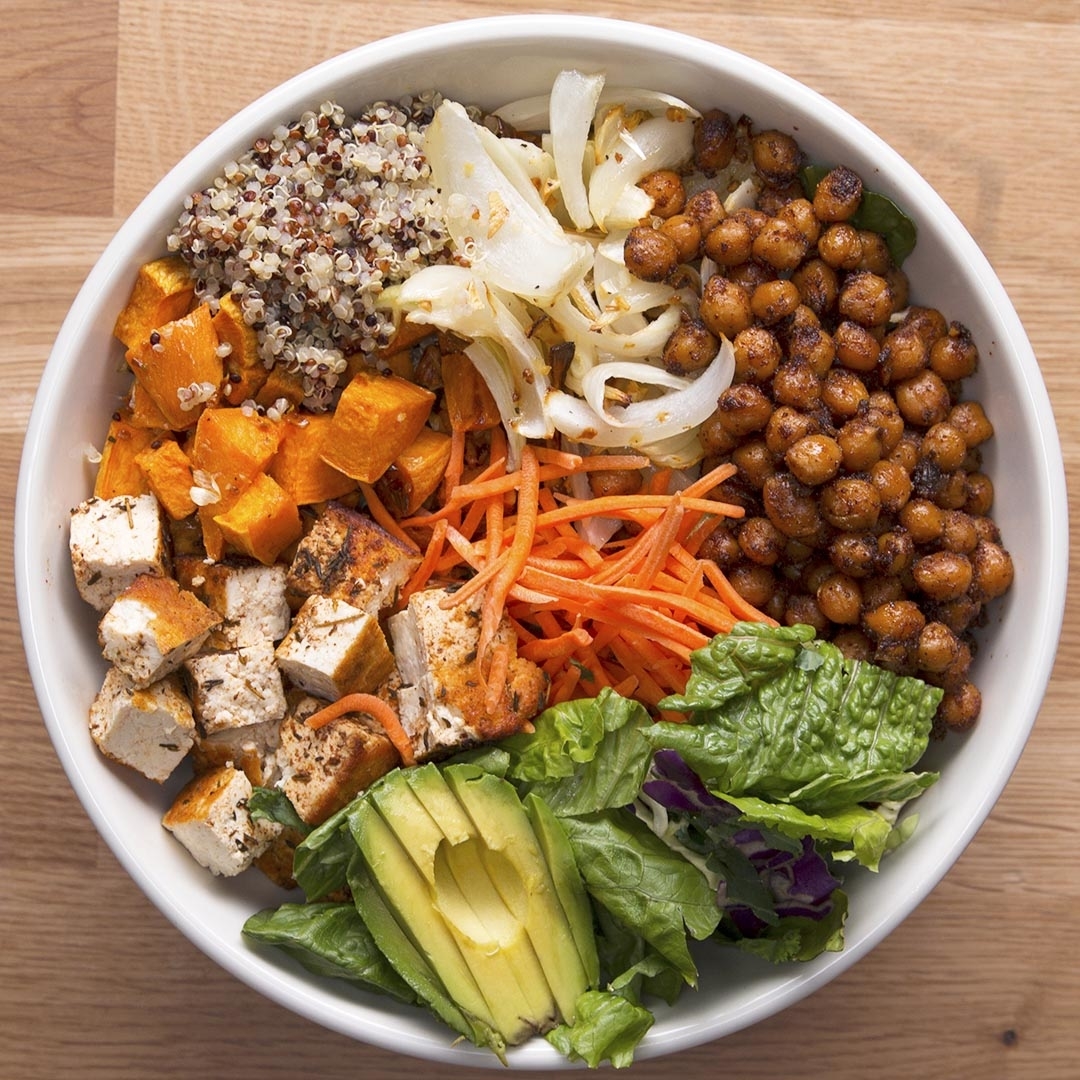 A bowl filled with quinoa, roasted chickpeas, sautéed onions, sweet potatoes, cubed tofu, shredded carrots, sliced avocado, and mixed greens
