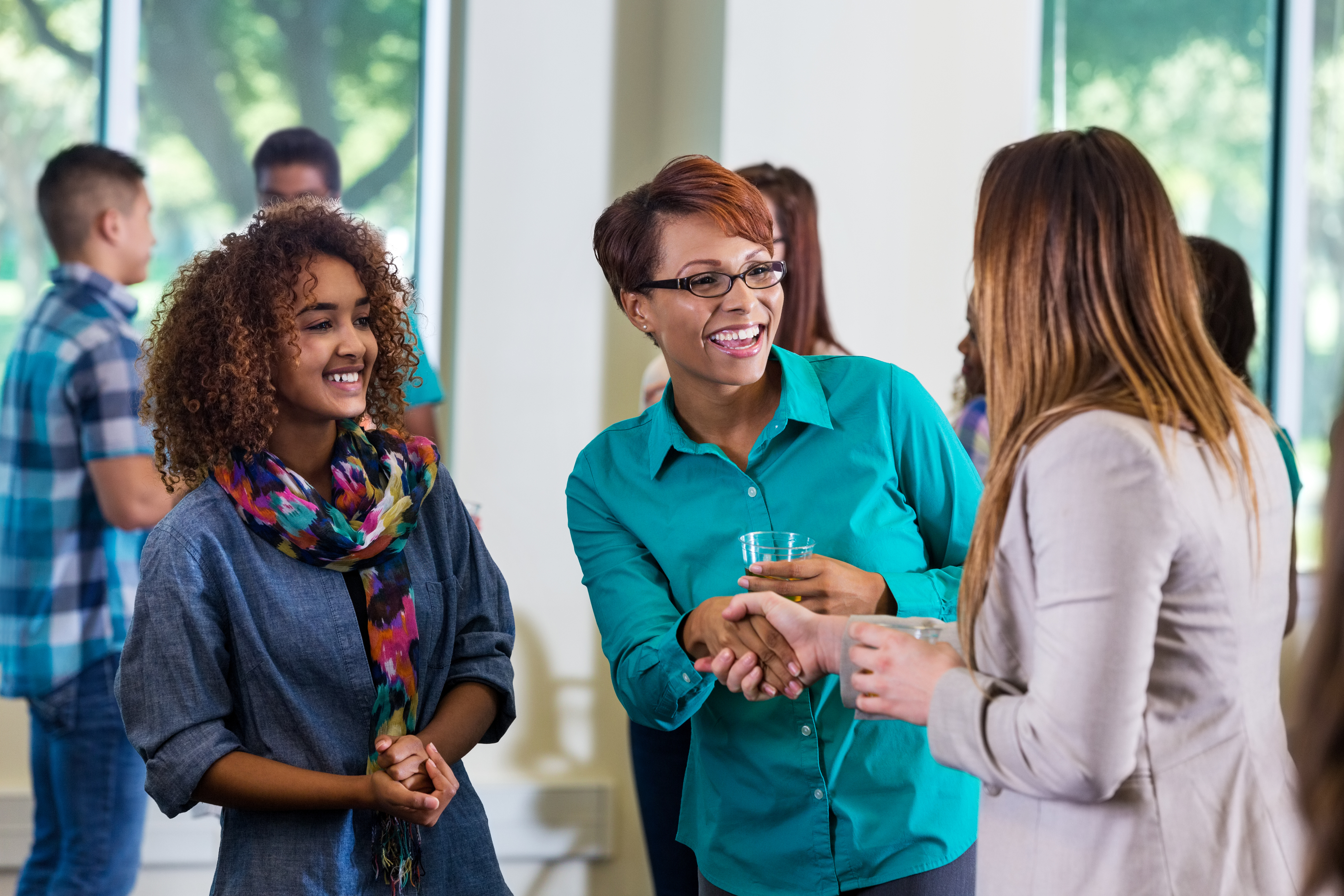 A group of people are mingling at a social event. Three women in the forefront are talking and smiling, one wearing a colorful scarf and another with a turquoise shirt. Names unknown