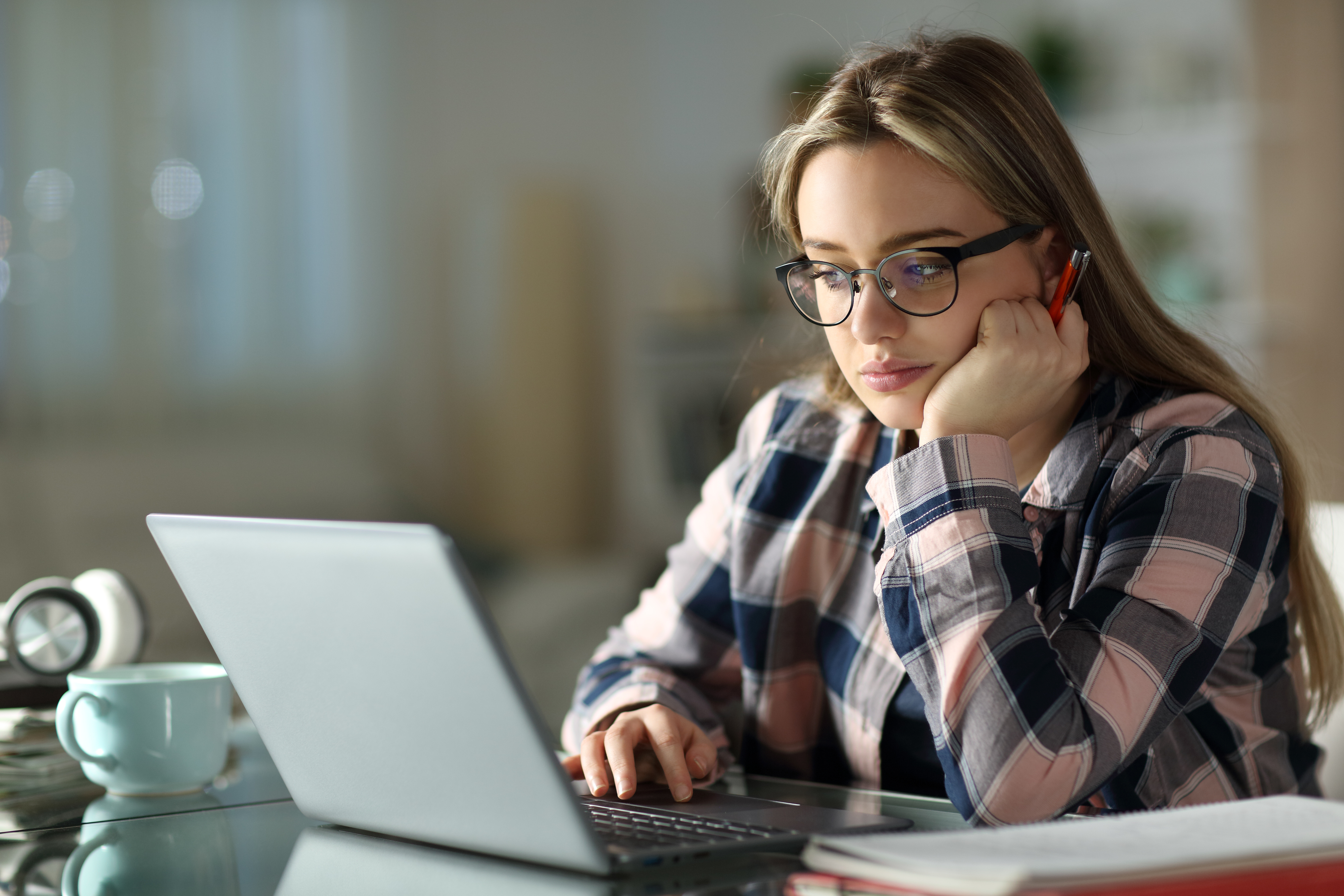 A person wearing glasses and a plaid shirt is sitting at a desk, looking at a laptop screen, holding a pen, with a notebook and mug nearby