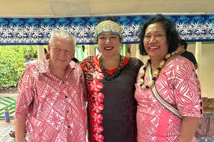 A man and two women smiling at a cultural event. The man and woman on the right are wearing floral patterns, while the woman in the center wears a traditional headdress