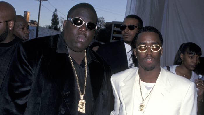 The Notorious B.I.G. and Sean Combs pose together at an event, both wearing sunglasses and gold chains, with The Notorious B.I.G. in a dark suit and Sean in a white suit