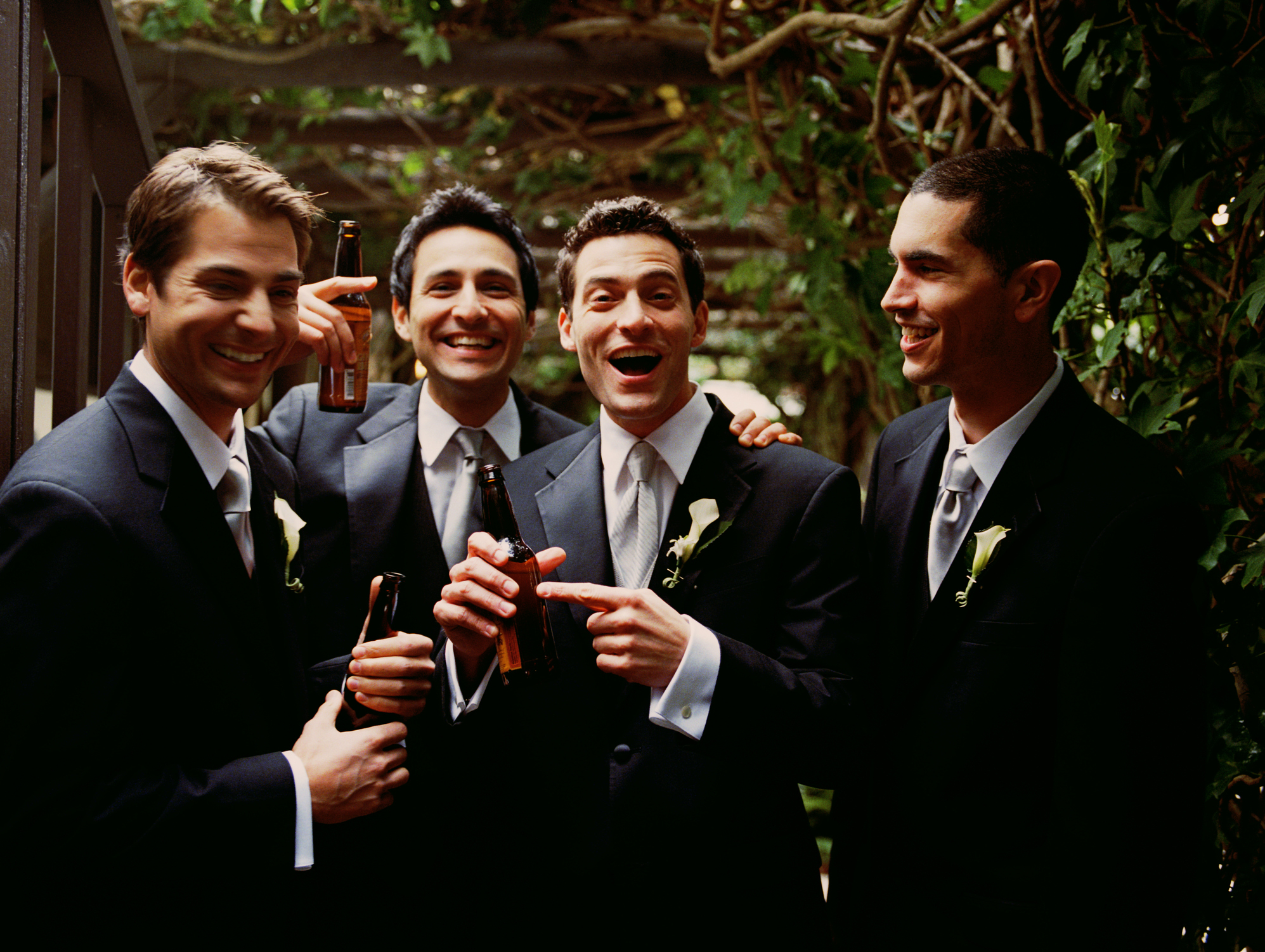 Four men in tuxedos, smiling and holding beer bottles, stand together in an outdoor setting. Names of the men are unknown