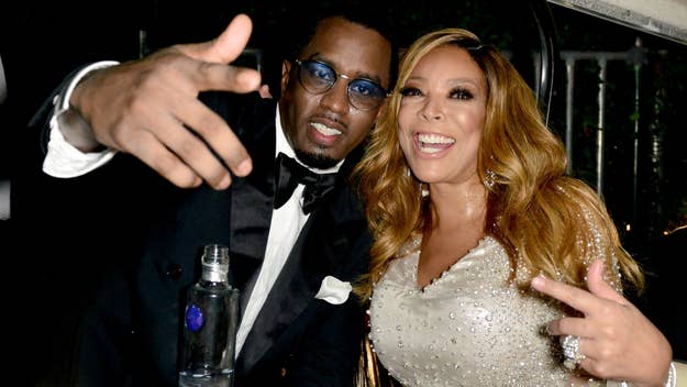 Diddy wearing a black tuxedo and Wendy Williams in a sparkling dress smile and gesture playfully at the camera