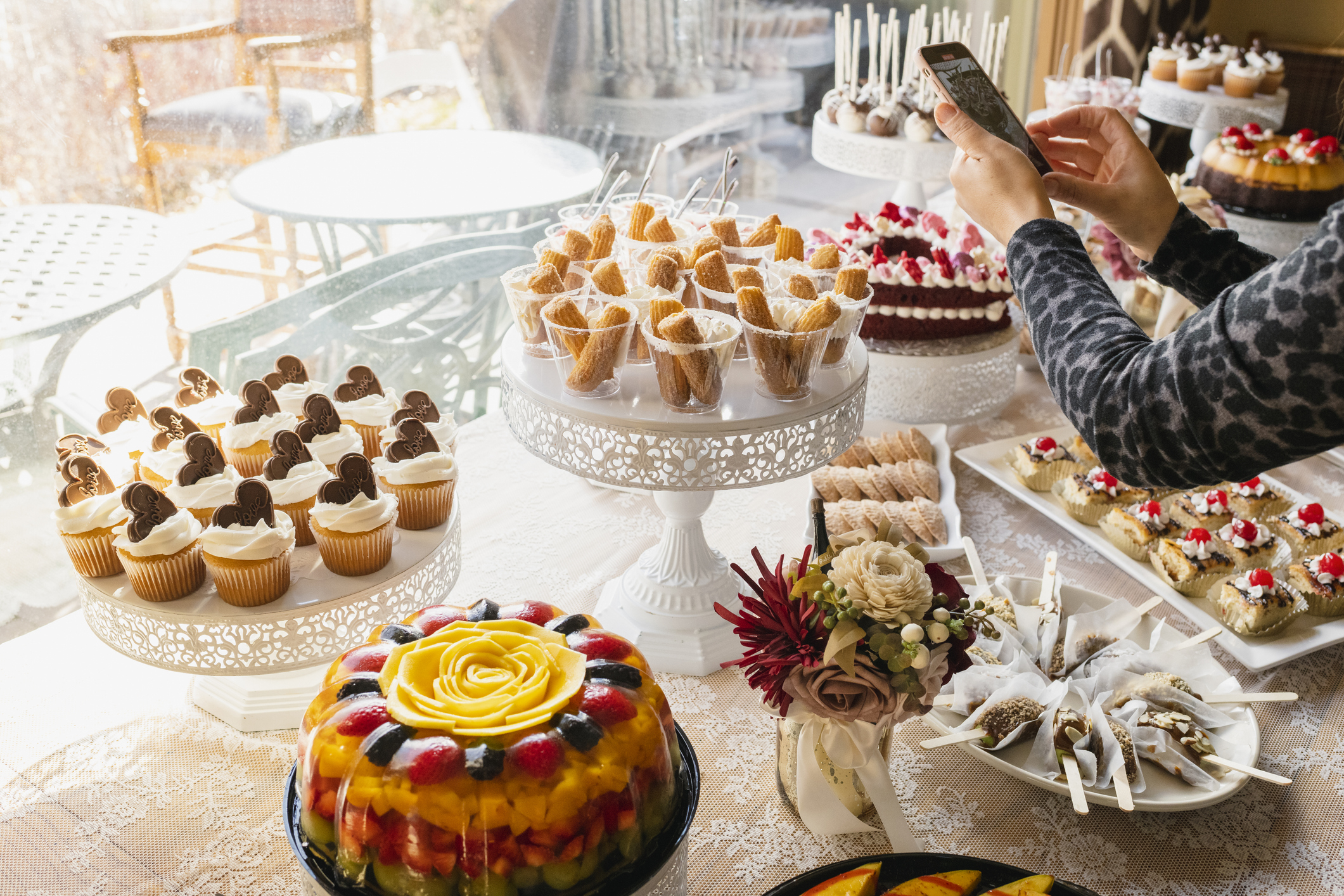 Assorted desserts, including cupcakes, fruit-topped cakes, and pastries, on a beautifully arranged table. A person takes a photo of the display with a smartphone