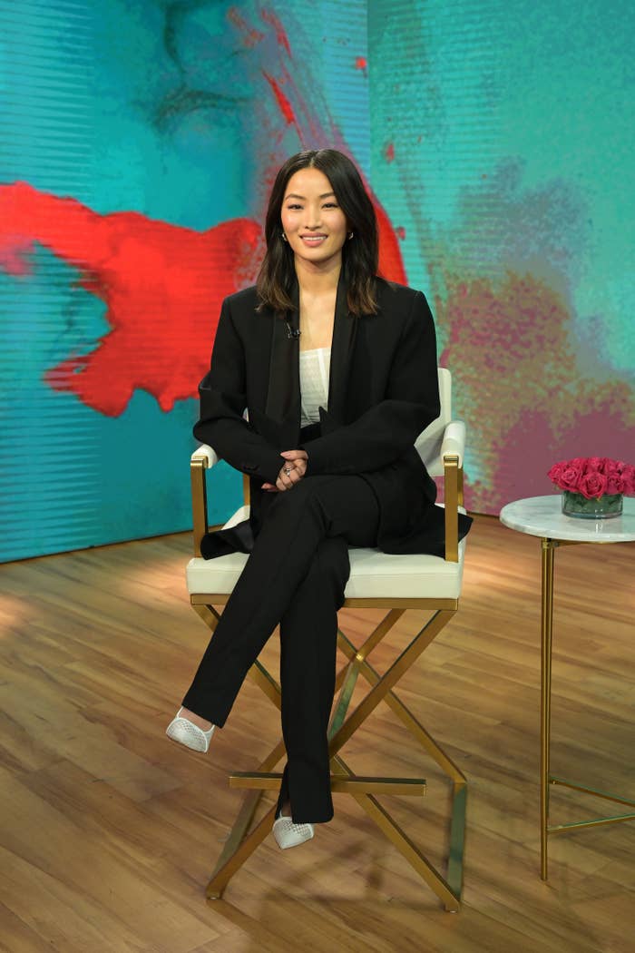 Anna Sawai, dressed in a tailored pantsuit, sits on a chair in a studio with a small table with flowers beside her