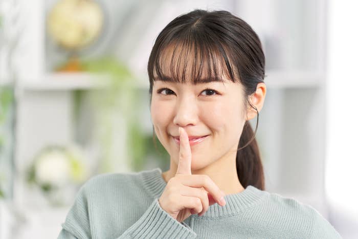 A woman with straight bangs and a light top smiles while holding a finger to her lips, suggesting silence